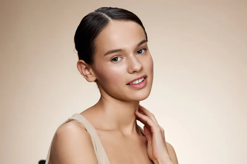 Young woman with natural makeup and neutral attire, showcasing a serene expression against a soft beige background, ideal for beauty and wellness concepts