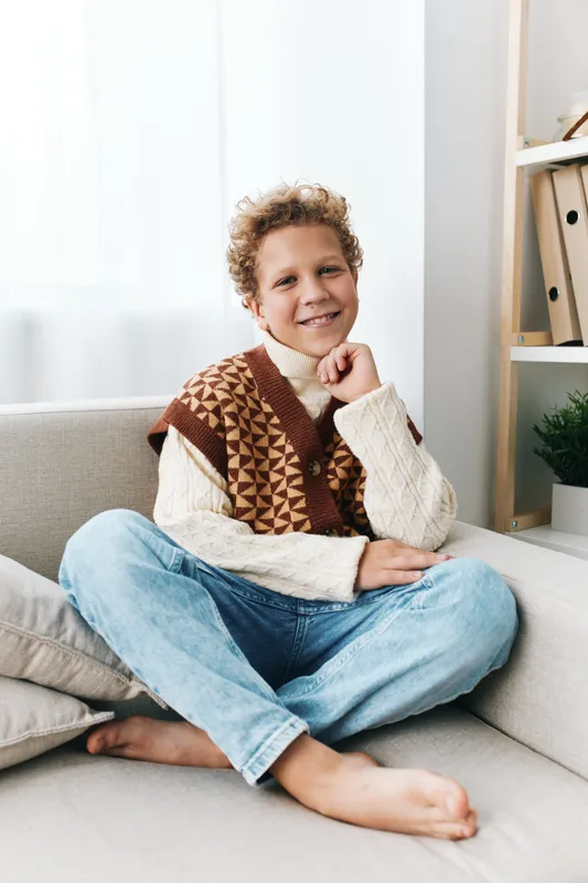 Playful young boy sits barefoot on a cozy sofa, wearing a knitted sweater and denim jeans, smiling warmly at the camera with relaxed casual charm.