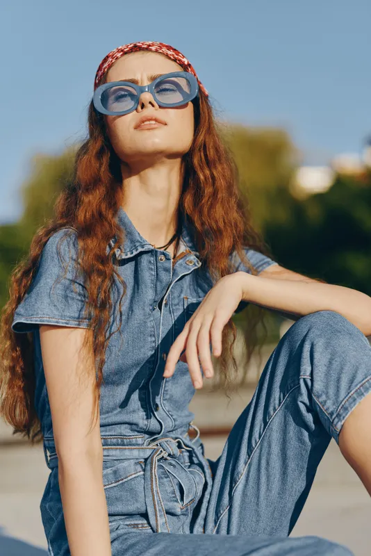 young woman in denim jumpsuit wearing blue sunglasses outdoors under sunlight with relaxed confident pose and natural background during daytime
