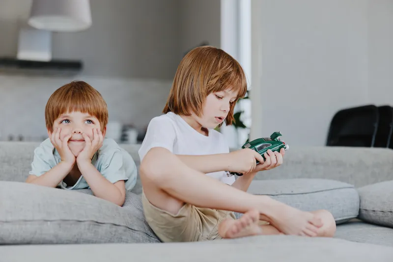 Two young boys engaging in play, one focused on a toy car while the other looks intrigued, creating a joyful atmosphere in a cozy living room.