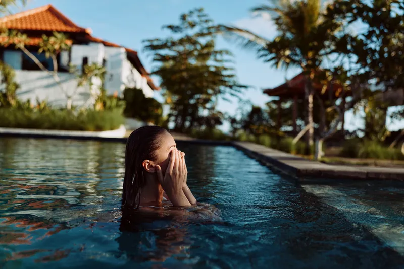 Person submerged in a pool, hair wet, hands shielding eyes, beside a resort villa under a blue sky. Lifestyle scene focused on relaxation, leisure, and warm water.