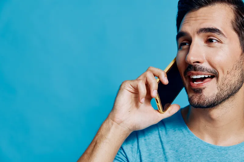 Happy smiling man chatting on mobile phone, wearing blue shirt, with a joyful expression on his face. Bright turquoise background adds energetic vibe to the scene.
