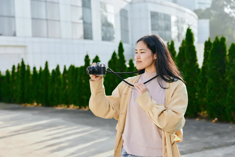 Candid photography scene of a young woman holding a camera, outdoors near modern buildings, casual attire, focused on framing a shot, daylight and calm urban atmosphere