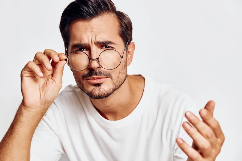 Curious and thoughtful young man wearing glasses looking intently at the camera, gesturing with his hand, isolated on a solid light background. Concept: inquiry and reflection.