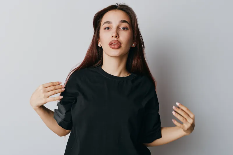 Young woman with long hair wearing black shirt making funny face with tongue out in studio against white background. Casual style, playful expression and relaxed pose for lifestyle portrait.