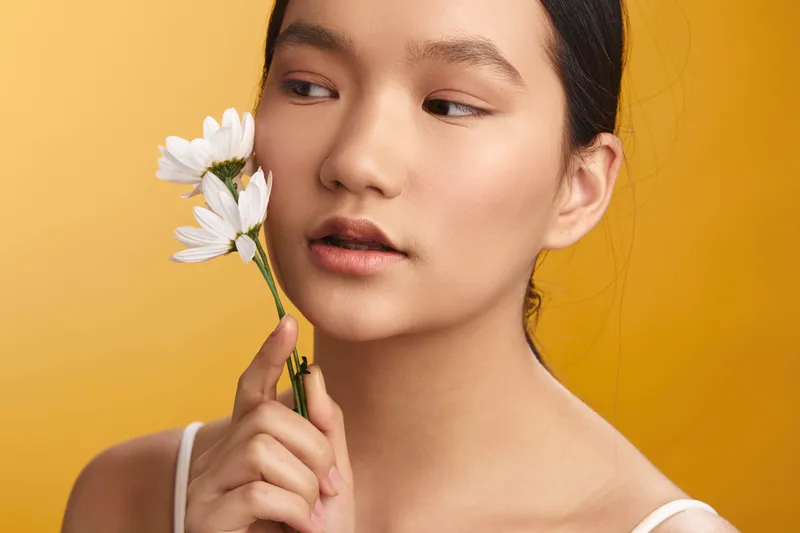 asian woman holds a delicate flower near her face, set against a warm yellow backdrop, conveying calm beauty and natural charm in a studio portrait.