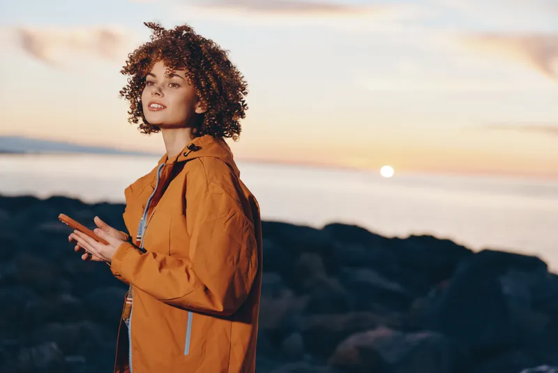 Woman lifestyle portrait outdoors by ocean at sunset, curly hair, orange jacket, standing and smiling, travel moment near coast, warm golden light, evening breeze