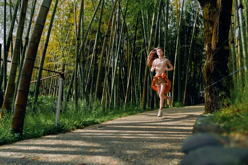 woman running bamboo forest path fitness outdoors motion joyful young female jogging on shaded gravel trail through tall bamboo grove summer workout active lifestyle and nature