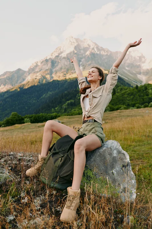 Happy woman in casual clothes sitting on a rock with arms raised in a mountain field. Concept of lifestyle, freedom, nature, and outdoor adventure. Bright natural light.