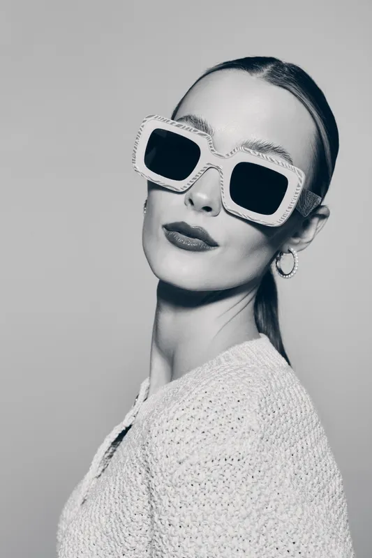 Caucasian woman with fair skin wearing large white sunglasses and knitted sweater poses in studio. Fashion and style concept.