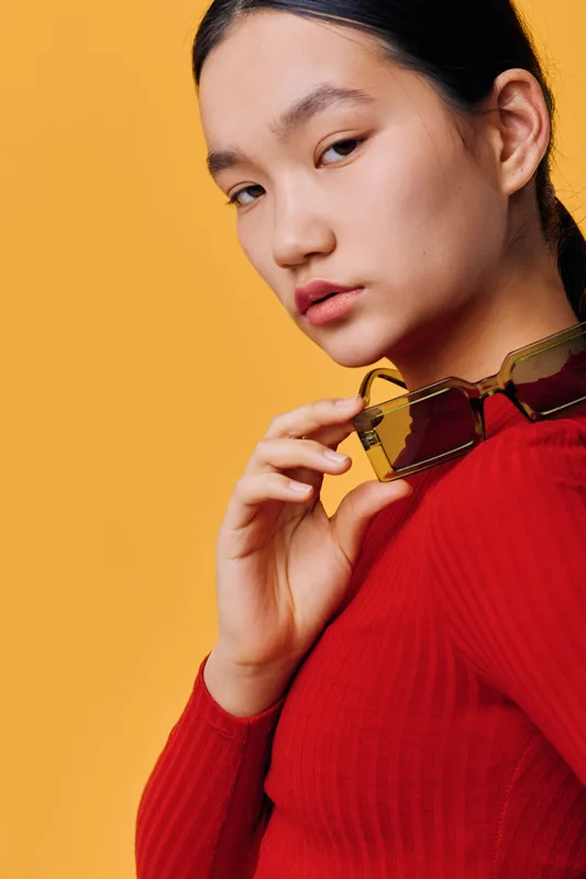 Young woman in red sweater holding sunglasses with elegant pose and neutral expression on yellow background. Fashion portrait with confident look and casual style for lifestyle and beauty.