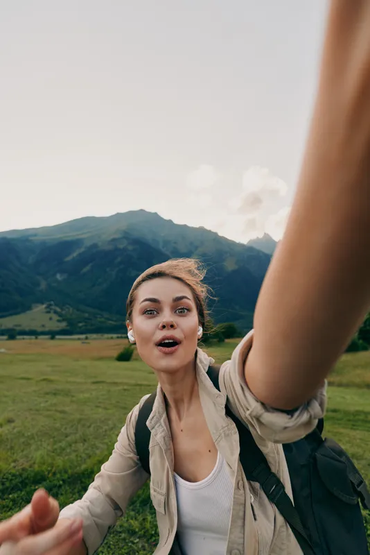 Young woman with backpack takes a selfie outdoors in a green meadow with mountains in the background, expressing excitement and adventure on a clear day.