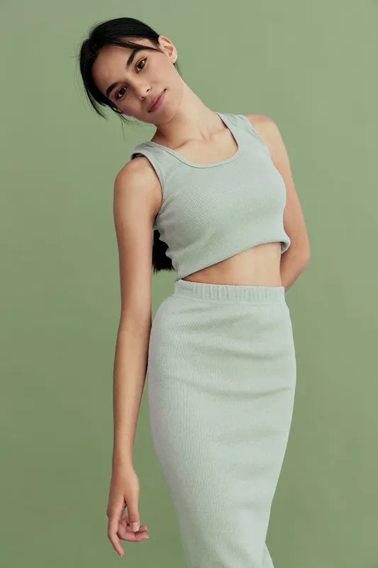 Graceful woman in light gray sleeveless crop top and matching skirt posing against pastel green background, showcasing minimalist fashion and soft natural lighting.