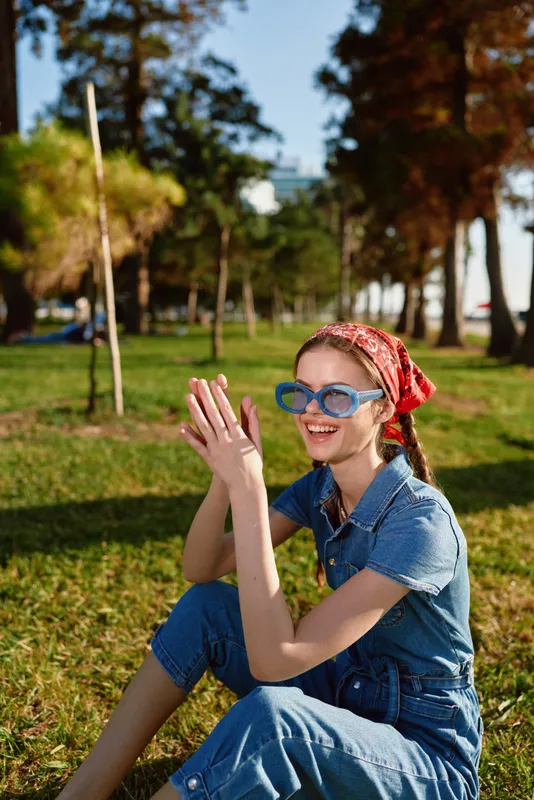Lifestyle woman in modern boho-western denim street style wearing blue sunglasses and a red bandana, enjoying sunny outdoor park with film color effect.