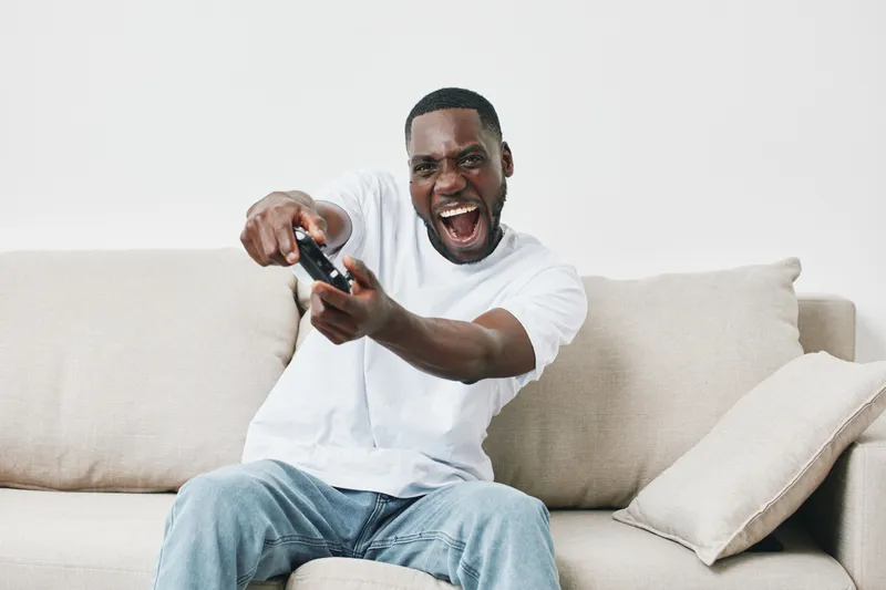 Excited young man playing video games at home, wearing a casual white t shirt and jeans, enjoying leisure time on a couch, vibrant atmosphere