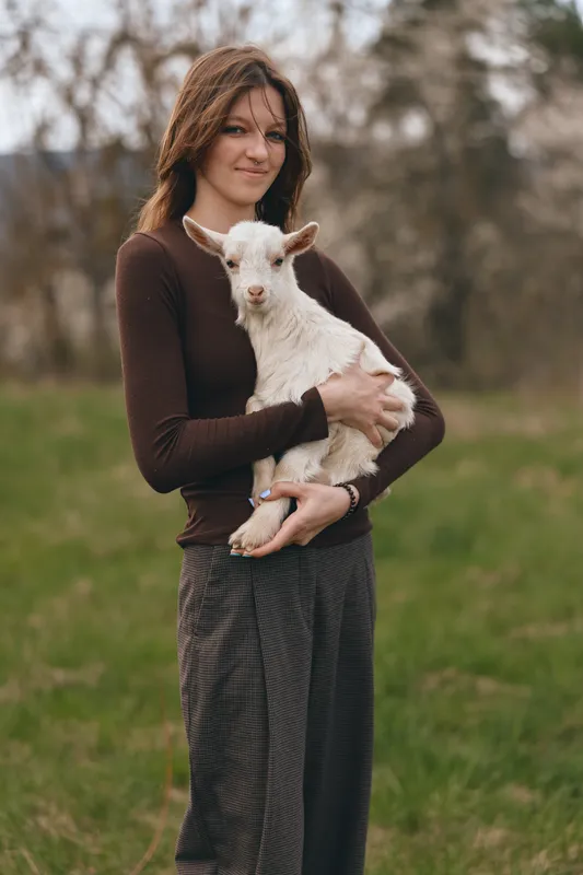 A woman in a brown sweater holds a young goat in an open field, smiling softly. An earthy rural portrait celebrating animal companionship, farming life, and gentle bonds between humans and livestock.