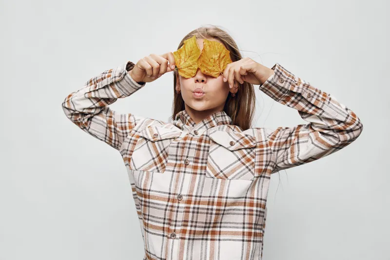 Girl in plaid shirt holding yellow autumn leaves over her eyes with playful expression, standing against plain gray background for seasonal fall concept.
