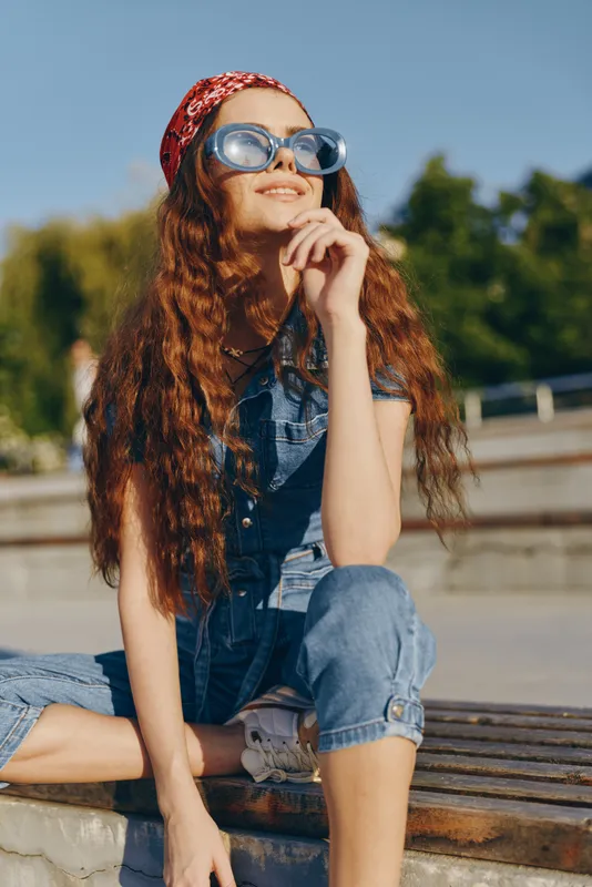 lifestyle woman in modern boho-western denim street style wearing blue sunglasses and a red bandana, sitting outdoors enjoying sunny weather with film color tones.