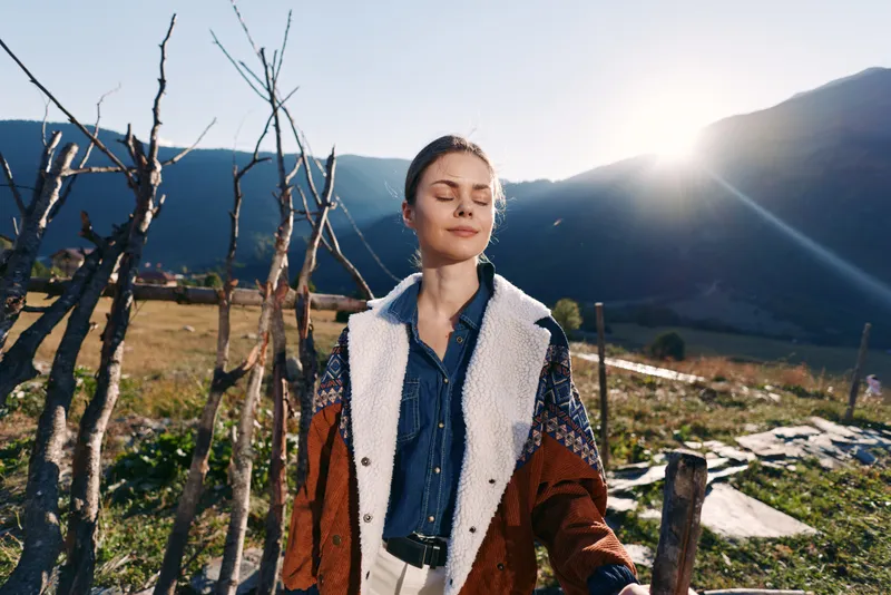 Woman portrait in mountains with sunlight and nature, peaceful expression with eyes closed, wearing warm jacket in rural outdoor scene, relaxation, fresh air and mindful moment.