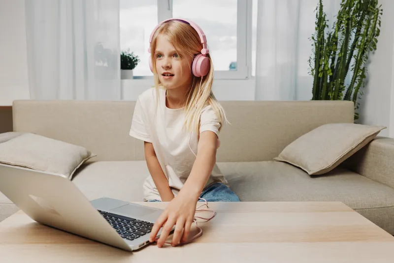 Young girl with pink headphones uses laptop at home sitting on sofa, engaged in technology and entertainment concept in bright living room interior.
