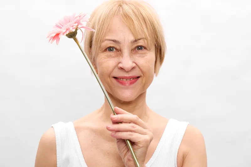 Woman portrait smiling elderly flower pink white closeup beauty, holding a thin stem with one blossom, soft studio background, calm wellness and gentle emotions