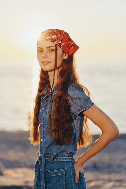 Young woman with long hair wearing a red bandana and denim jumpsuit stands by the sea at sunset, with soft sunlight creating a warm and relaxed atmosphere.