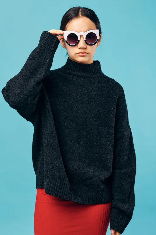 Woman wearing black sweater and oversized round sunglasses posing in studio with blue background. Casual style with confident look and modern fashion outfit for autumn or winter season.