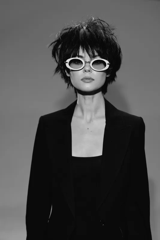 Black and white portrait of a stylish woman wearing white sunglasses and a black blazer with short messy hair.