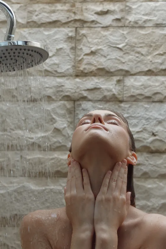 woman taking shower with water running on face and neck, relaxing under indoor shower, refreshing skincare moment in bathroom setting