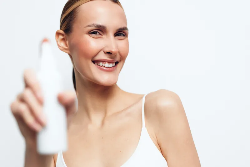 Young woman with blonde hair smiles while holding a white cosmetic bottle in a bright studio setting wearing a white tank top, expressing confidence and freshness.