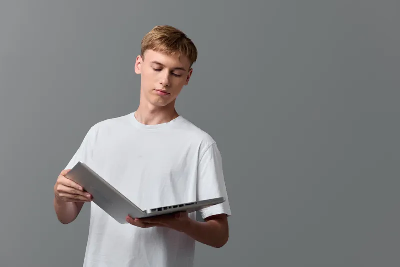 Casual young man reading on a laptop, focused expression, simple studio backdrop, modern technology at hand, learning mindset and relaxed pose for education or work scenarios