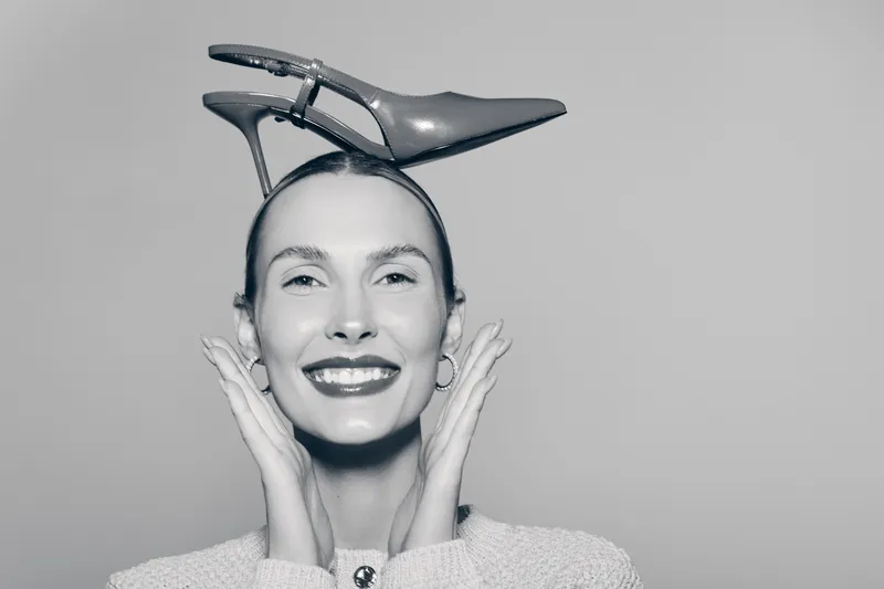 Smiling caucasian woman with light skin and slicked hair holds a high heel shoe on her head, posing against a plain grey background. Concept for fashion, style, and playful portrait.