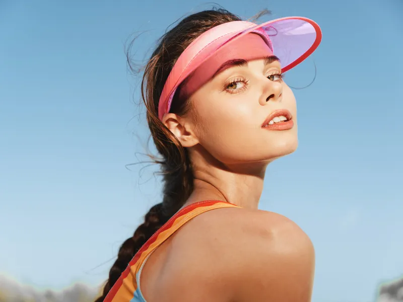 Portrait of a confident woman wearing a pink visor, sunlit skin and braided hair, set against a clear blue sky with warm summer light and a stylish athletic vibe.