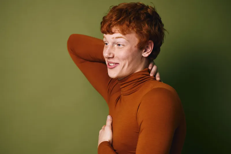 Young man with red hair wearing orange turtleneck sweater posing relaxed and smiling with hand behind head on green background in studio lighting portrait.