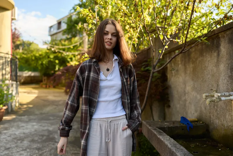 A young woman in a plaid shirt, white tee and gray sweatpants strolls along a sunlit garden alley, exuding casual confidence and a relaxed, carefree mood