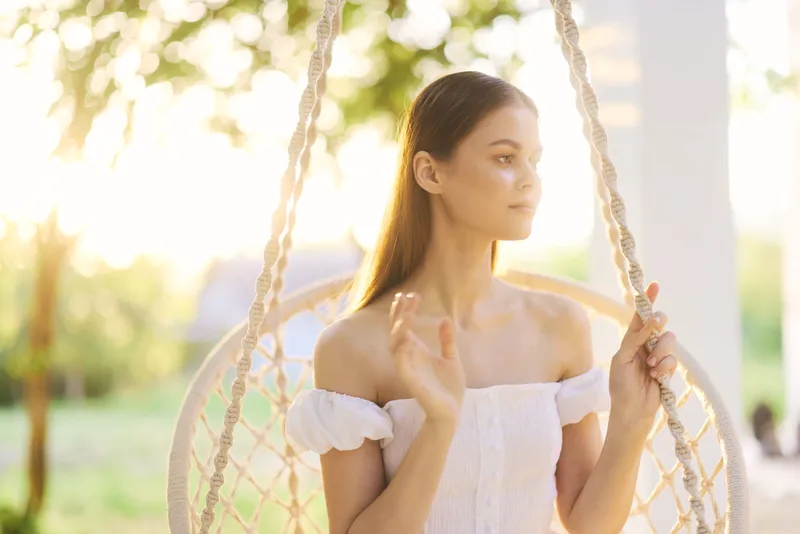 Young woman in white dress sitting on a swing, experiencing a moment of calm in a sunlit garden, showcasing peace and tranquility with nature