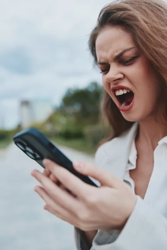frustrated woman holding smartphone outdoors showing anger facial expression with natural background and casual jacket in emotional moment financial crisis concept