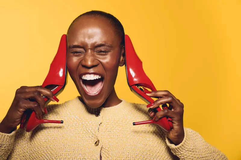 Happy dark skinned African woman with short hair holding red high heels near ears, laughing with eyes closed on yellow background. Concept of joy, fashion, and vibrant lifestyle.