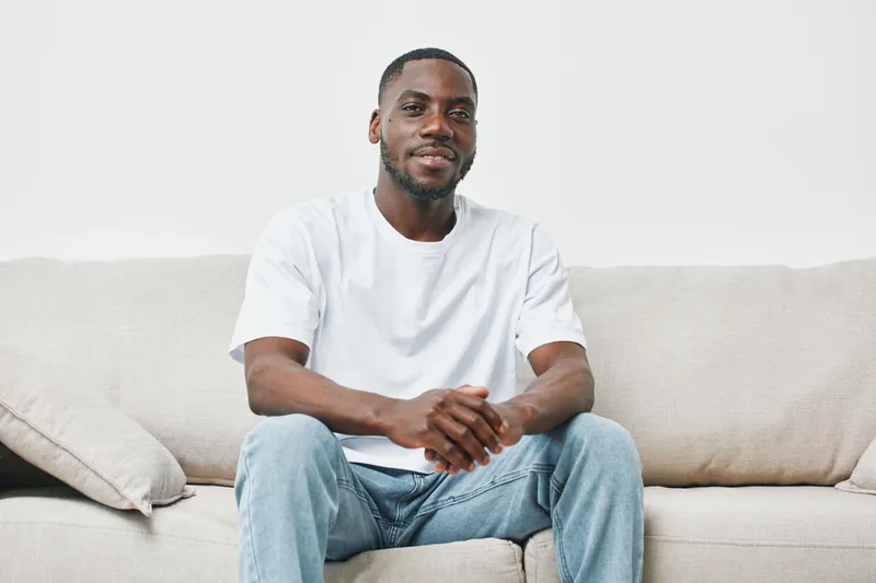 Smiling young African American man sitting on a couch in casual attire, representing comfort and relaxation in a modern home setting