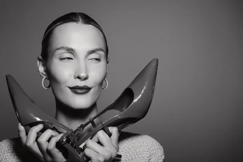 White woman with fair skin holding shiny high heel shoes near face with closed eyes. Concept of fashion accessories, style, and elegance in monochrome studio.