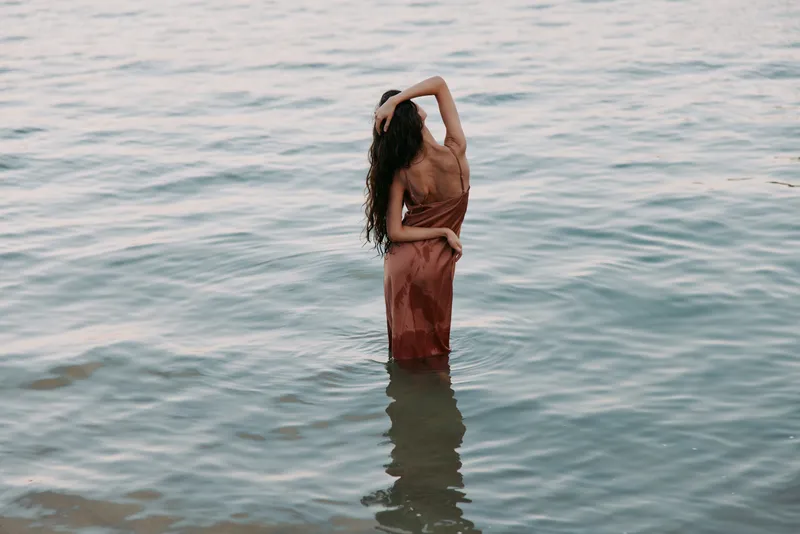 A lone figure stands waist-deep in calm water, back turned, wearing a flowing dress as sunlight glints on gentle ripples.