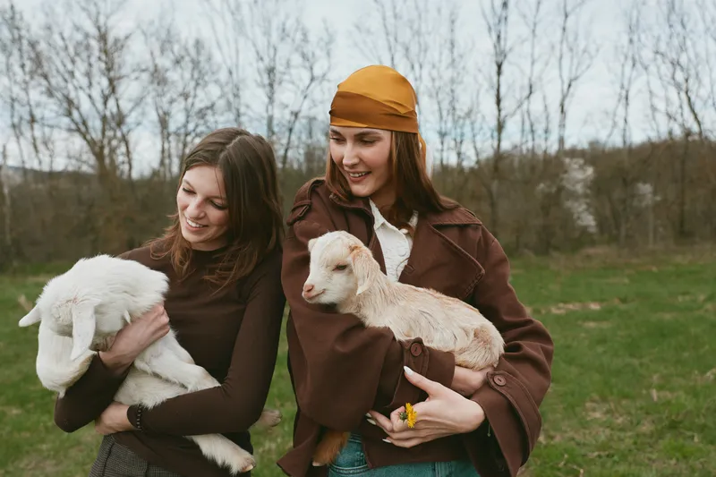 Two women outdoors cradle young goats, sharing a tender moment in a field. Casual countryside lifestyle with friendly animals and natural bonding. Soft daylight and earthy tones create a warm,