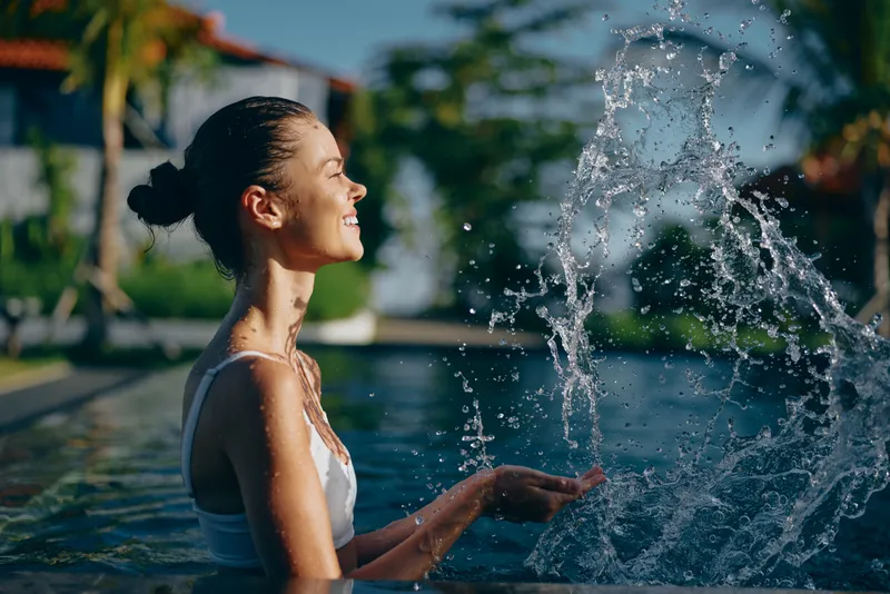 A smiling woman enjoys a playful splash by a sunny pool, savoring leisure and relaxation as water arcs from a fountain. Concept for outdoor lifestyle and carefree summer moments.