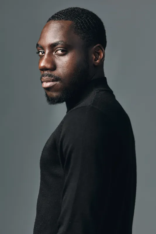 African man portrait in creative conceptual visual style with dark skin and black sweater looking sideways on gray background. Studio lighting highlights facial features and expression.