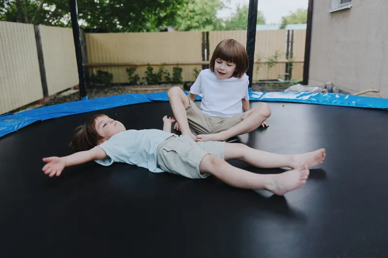 Two cheerful children playing on a trampoline in a backyard, with one boy lying down and the other sitting and laughing joyfully. Perfect for childhood fun and outdoor activities.