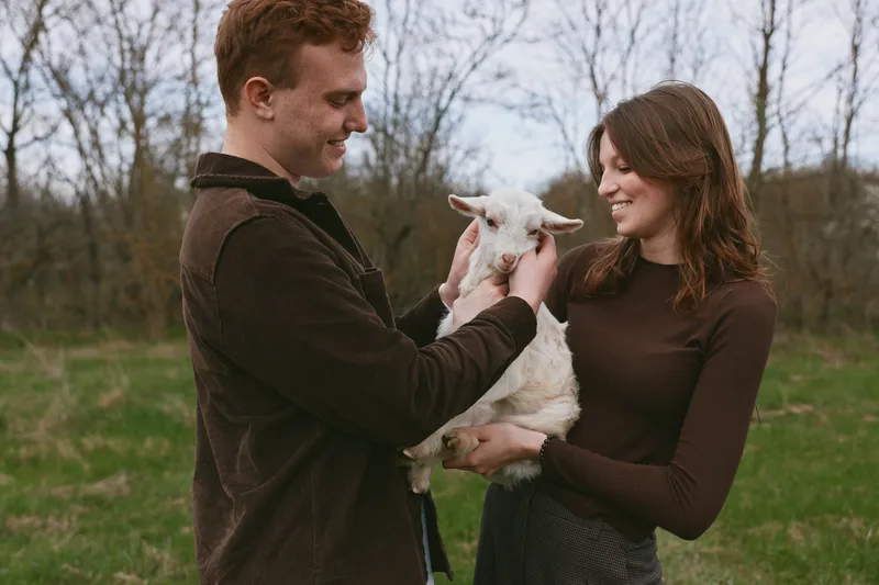 Smiling couple outdoors gently holds a young goat kid in a grassy countryside field, creating a wholesome lifestyle moment on a farm. Natural light, relaxed pose, and friendly, affectionate