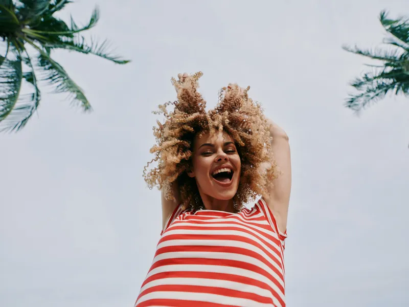Joyful woman with curly hair and striped red and white top raises her arms in the sunny sky, capturing a moment of carefree energy and summer vitality.