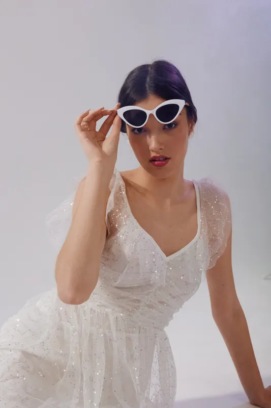 Young woman in a white sparkling dress wearing white cat-eye sunglasses. She poses confidently in a studio with a neutral background. Concept of woman beauty fashion.