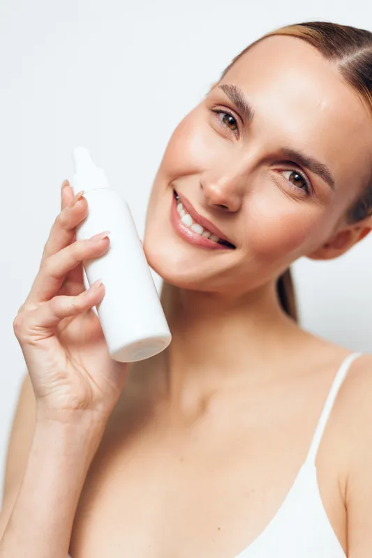 Young woman holds a skincare serum bottle and applies it with a dropper on her face, smiling against a clean white background. Beauty routine selfcare.