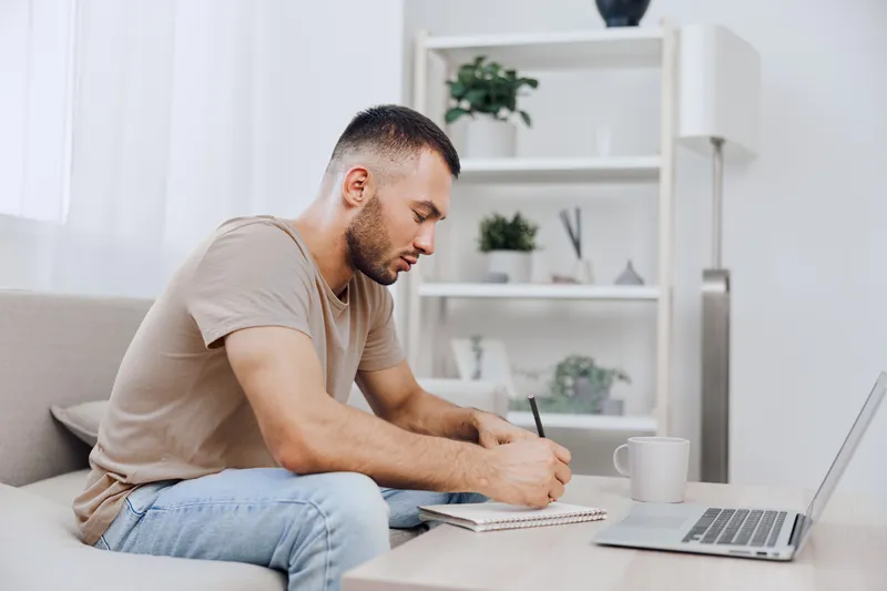 Focused man writing notes while sitting on a cozy couch, surrounded by a bright and modern interior, capturing the essence of productivity and creativity in a contemporary home.